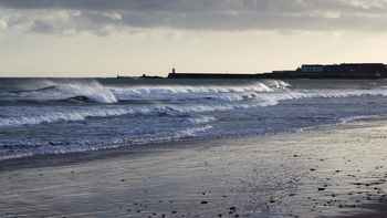 Seahouses waves The image is a landscape photograph showing waves breaking on the beach at Seahouses, located on the Northumberland coast in England, United Kingdom. In the far distance, buildings and the harbour wall of Seahouses are visible, with the sea stretching towards Bamburgh. The scene was captured in the morning during the winter season, as seen by the low sun and the subdued winter lighting. The main subject of the image is the dynamic movement of the waves on the sea, with the shoreline of Seahouses beach in the foreground. The photograph highlights the natural beauty of the Northumberland coast, with the town of Seahouses providing a coastal backdrop.
